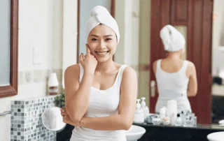 Asian woman in bathroom with white towel wrapped around head, face on hands and smiling